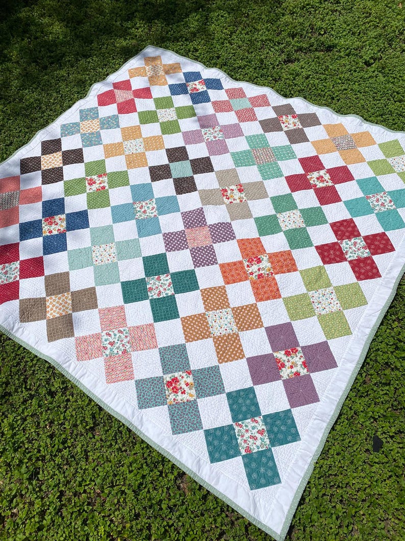 K&ouml;nnte beinhalten: Eine quadratische Patchwork-Decke mit einem sich wiederholenden Kreuzmuster aus verschiedenen bunten Stoffquadraten. Das Design umfasst Blumen-, Punkte- und Unimuster in Rot-, Blau-, Gr&uuml;n-, Orange- und Violettt&ouml;nen auf wei&szlig;em Hintergrund mit einem hellgr&uuml;nen Rand.