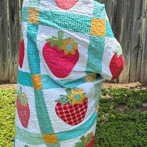 May include: A quilted blanket featuring a repeating pattern of red strawberry appliqu&eacute;s with green leaves and yellow flowers. The quilt is constructed with white blocks separated by teal and yellow grid lines, all finished with intricate swirling stitching.
