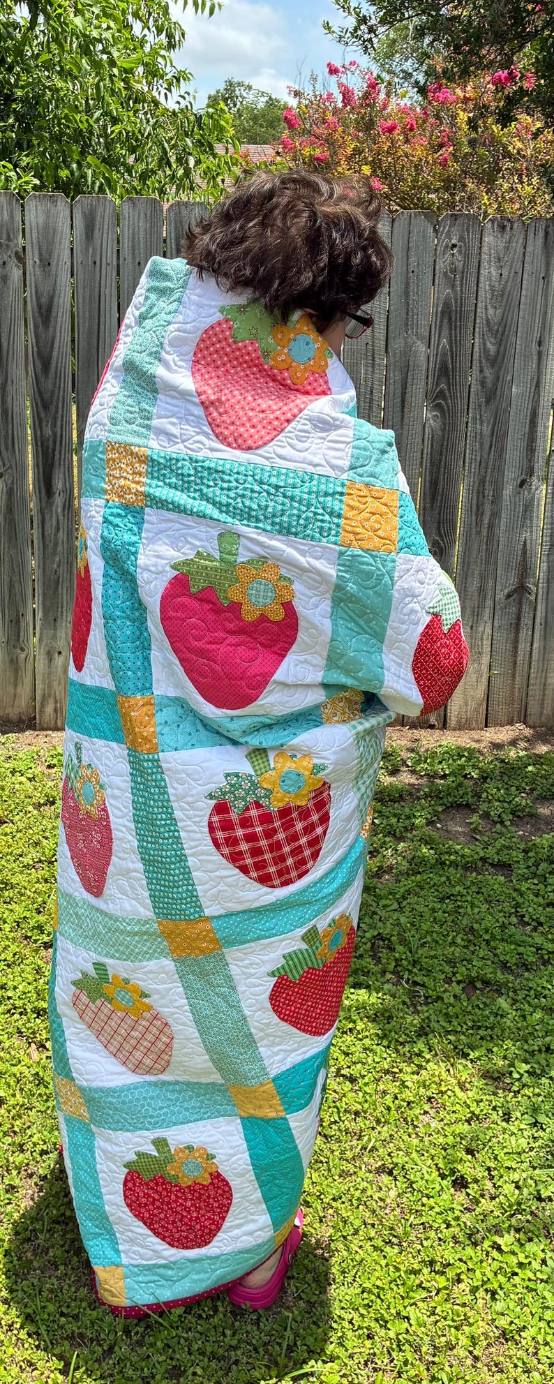 May include: A quilted blanket featuring a repeating pattern of red strawberry appliqu&eacute;s with green leaves and yellow flowers. The quilt is constructed with white blocks separated by teal and yellow grid lines, all finished with intricate swirling stitching.