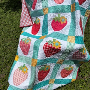 May include: A patchwork quilt featuring a repeating pattern of red strawberry appliqu&eacute;s with yellow and blue flower accents. The quilt is constructed with white squares separated by teal, green, and yellow patterned fabric strips with quilted texture.