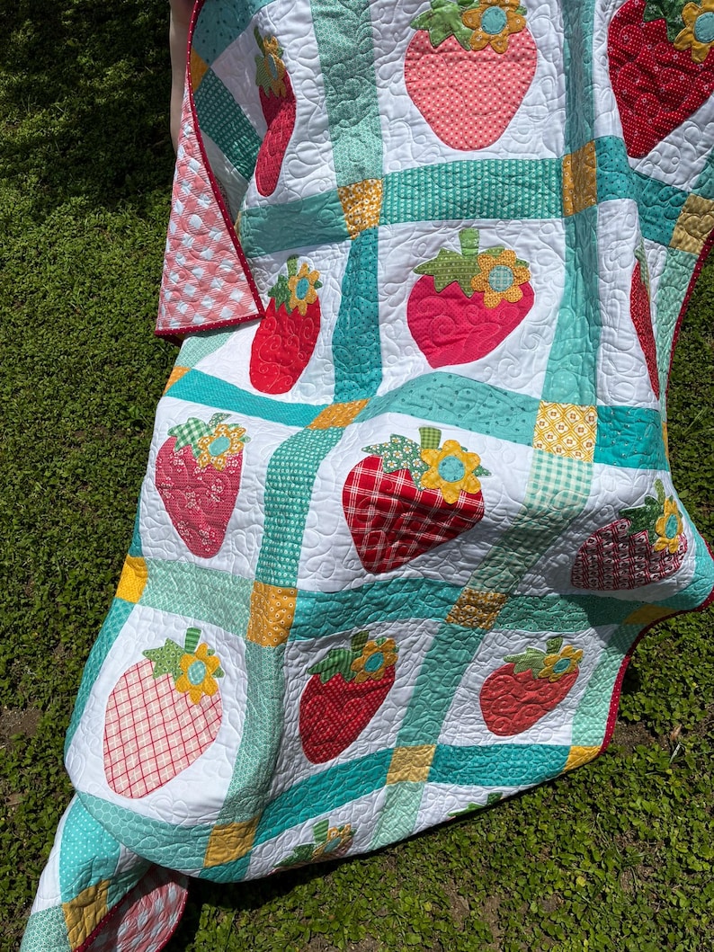 May include: A patchwork quilt featuring a repeating pattern of red strawberry appliqu&eacute;s with yellow and blue flower accents. The quilt is constructed with white squares separated by teal, green, and yellow patterned fabric strips with quilted texture.