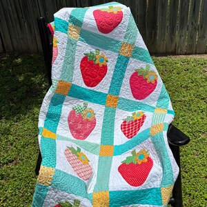 May include: A quilted blanket featuring a grid pattern of white squares with red strawberry appliqu&eacute;s and green leaves, separated by teal and yellow fabric strips. Each strawberry is topped with a small yellow and blue flower accent.