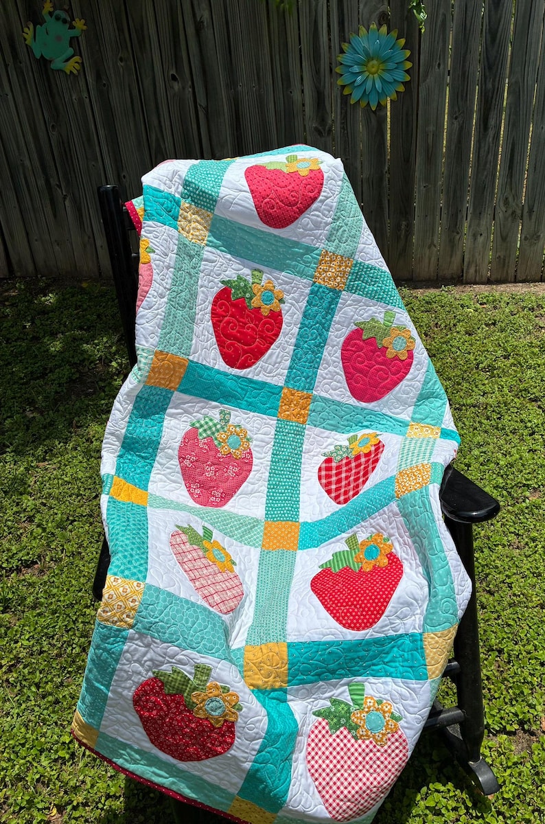 May include: A quilted blanket featuring a grid pattern of white squares with red strawberry appliqu&eacute;s and green leaves, separated by teal and yellow fabric strips. Each strawberry is topped with a small yellow and blue flower accent.