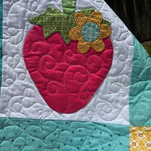 May include: A quilted fabric piece featuring a large pink strawberry appliqu&eacute; with a green stem and a yellow flower accent. The background is white with swirling stitch patterns, bordered by teal fabric sections with a subtle white dot pattern.