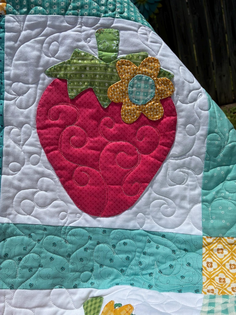 May include: A quilted fabric piece featuring a large pink strawberry appliqu&eacute; with a green stem and a yellow flower accent. The background is white with swirling stitch patterns, bordered by teal fabric sections with a subtle white dot pattern.