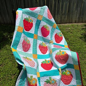May include: A patchwork quilt featuring a repeating pattern of red strawberries with green leaves and yellow flowers on a white background, separated by teal and yellow grid borders with a red polka dot fabric trim along the outer edge.