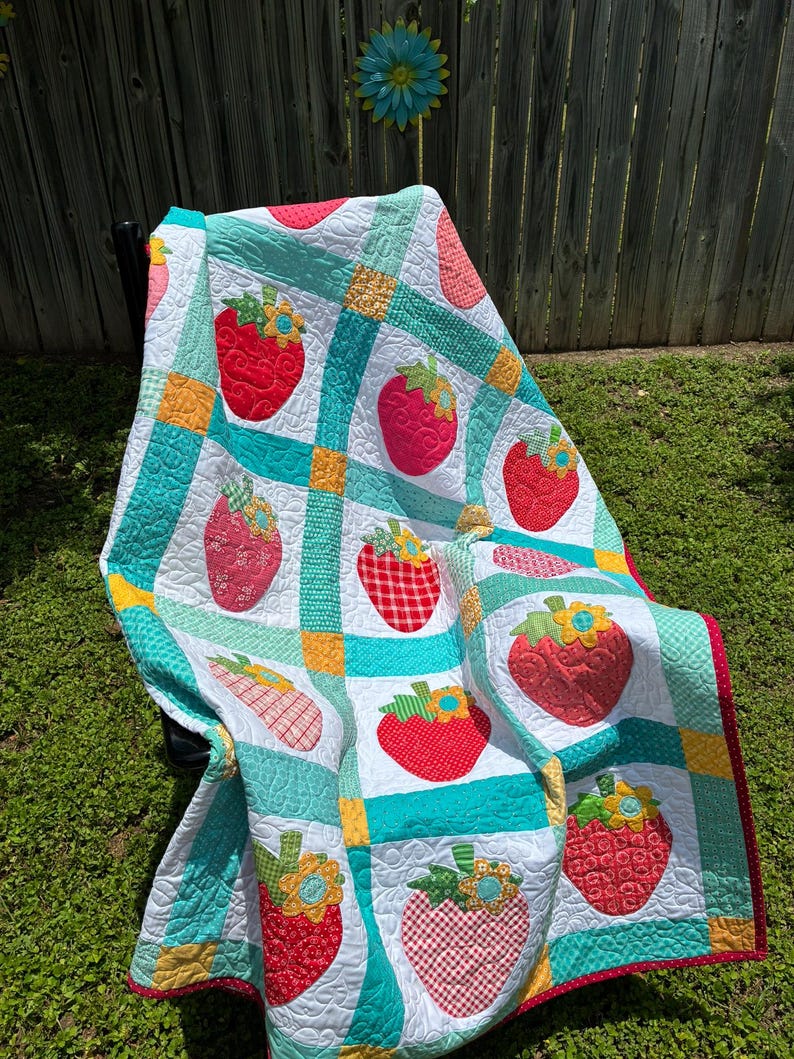 May include: A patchwork quilt featuring a repeating pattern of red strawberries with green leaves and yellow flowers on a white background, separated by teal and yellow grid borders with a red polka dot fabric trim along the outer edge.