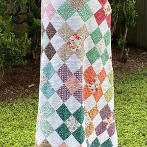 K&ouml;nnte beinhalten: Eine Patchwork-Decke mit einem sich wiederholenden Rautenmuster aus verschiedenen floralen und einfarbigen Stoffquadraten in den Farben Rosa, Gr&uuml;n, Orange, Braun und Wei&szlig;, wobei wei&szlig;e Stoffquadrate die farbigen, gemusterten Abschnitte trennen.