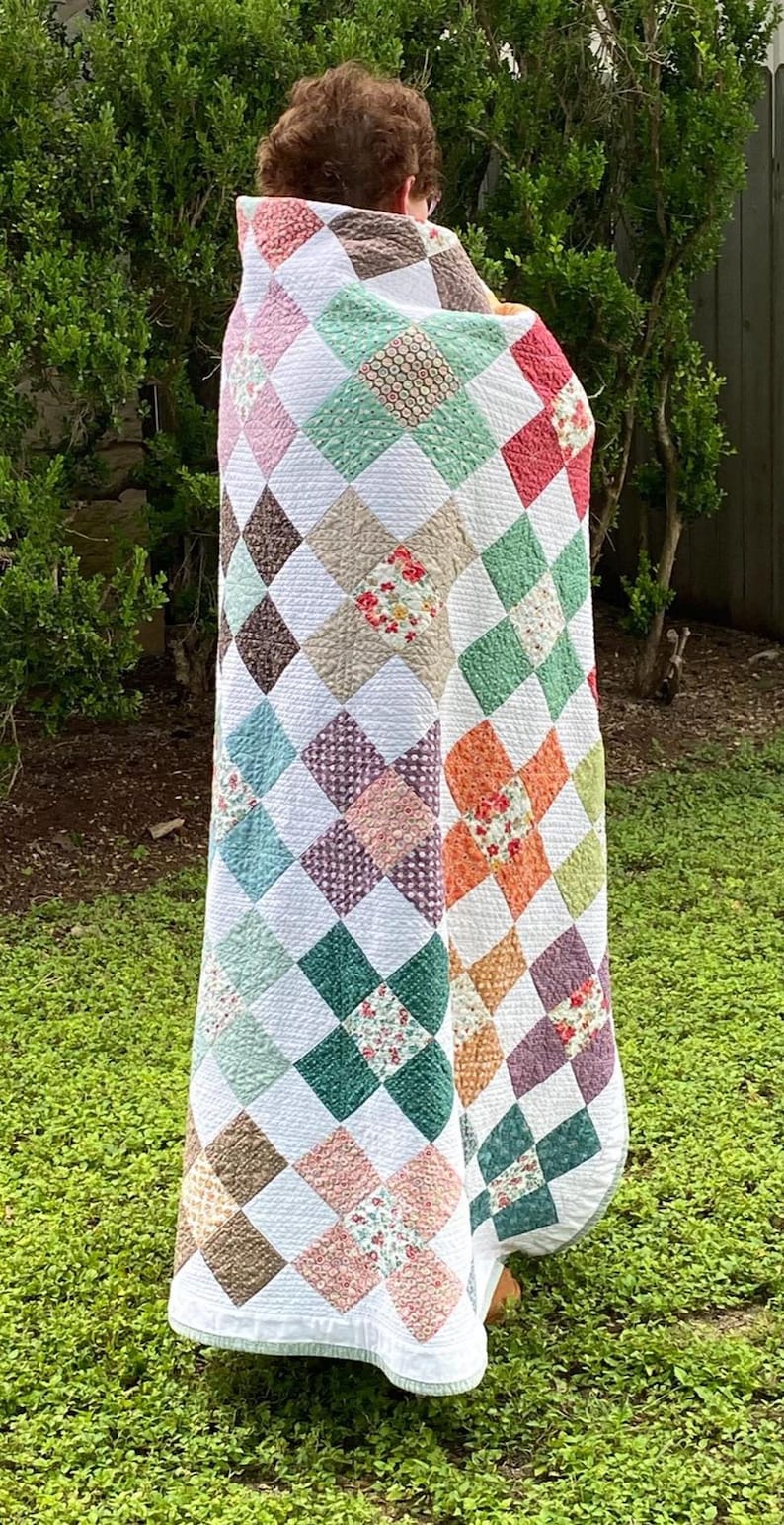 K&ouml;nnte beinhalten: Eine Patchwork-Decke mit einem sich wiederholenden Rautenmuster aus verschiedenen floralen und einfarbigen Stoffquadraten in den Farben Rosa, Gr&uuml;n, Orange, Braun und Wei&szlig;, wobei wei&szlig;e Stoffquadrate die farbigen, gemusterten Abschnitte trennen.
