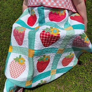 May include: A handmade quilt featuring a repeating pattern of red strawberries with yellow and blue flowers on a white background. The quilt is divided by teal and green checkered sashing with yellow corner squares and a pink gingham border.