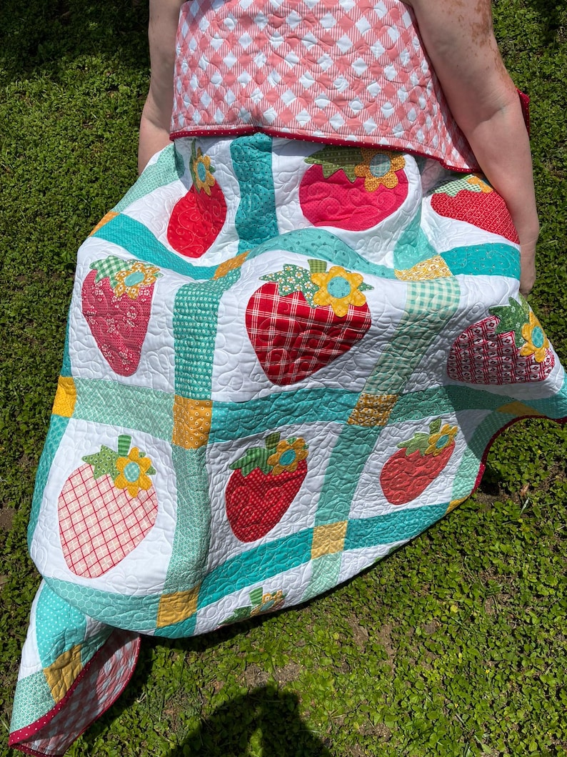 May include: A handmade quilt featuring a repeating pattern of red strawberries with yellow and blue flowers on a white background. The quilt is divided by teal and green checkered sashing with yellow corner squares and a pink gingham border.