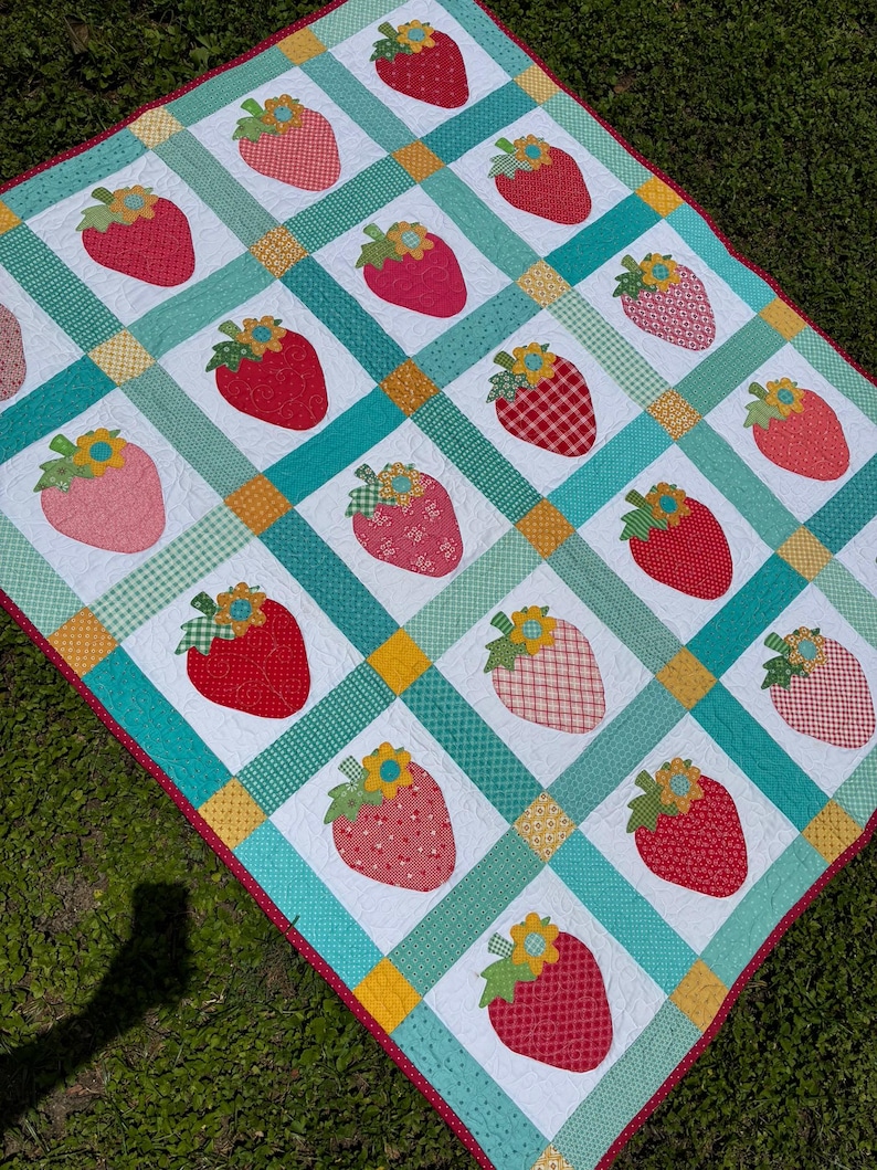 May include: A quilt featuring a grid of white squares, each displaying a strawberry appliqu&eacute; with a yellow flower and green leaves. The squares are separated by teal and yellow patterned fabric strips, finished with a red polka dot border.