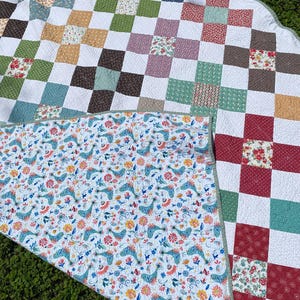 K&ouml;nnte beinhalten: Eine runde Steppdecke mit einem Patchwork-Design aus wei&szlig;en Quadraten, die sich mit Quadraten in verschiedenen Blumen- und geometrischen Mustern in Rot, Gr&uuml;n, Blau und Gelb abwechseln. Die Unterseite zeigt einen wei&szlig;en Stoff mit blauem Vogelmuster.