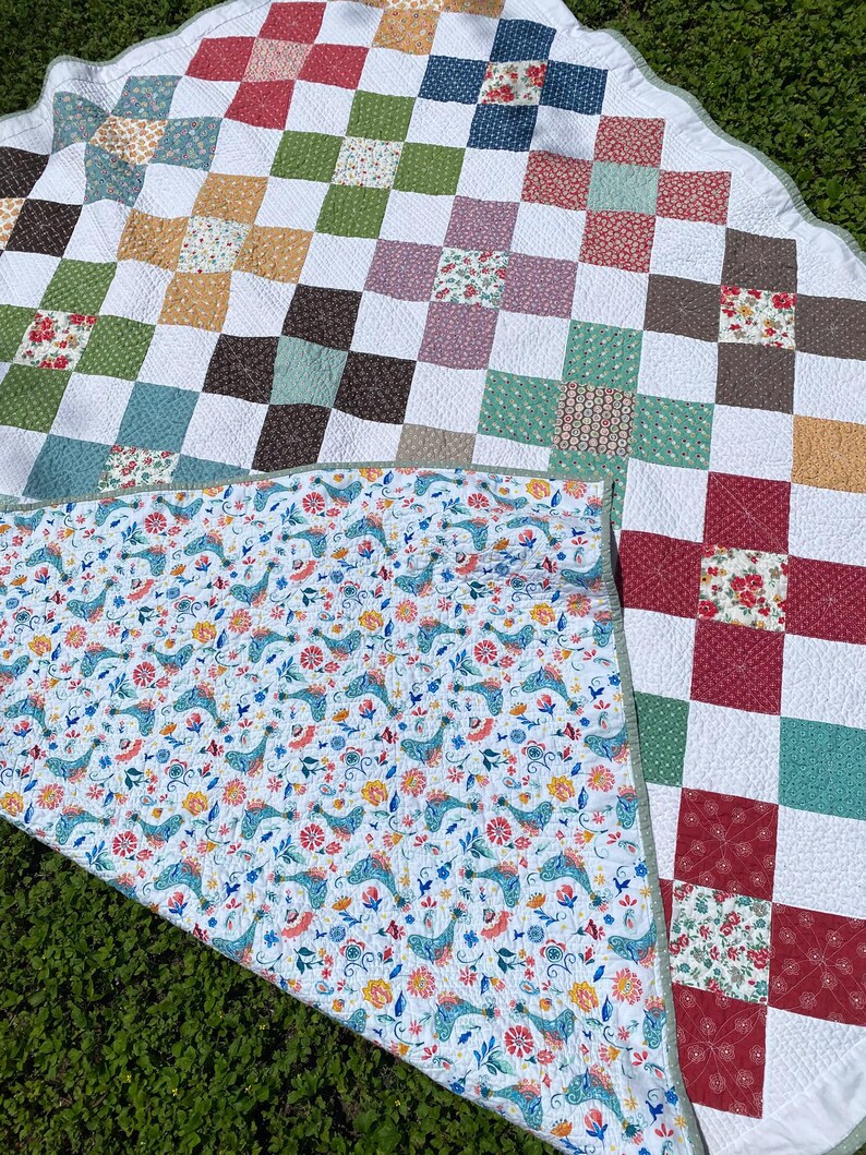 K&ouml;nnte beinhalten: Eine runde Steppdecke mit einem Patchwork-Design aus wei&szlig;en Quadraten, die sich mit Quadraten in verschiedenen Blumen- und geometrischen Mustern in Rot, Gr&uuml;n, Blau und Gelb abwechseln. Die Unterseite zeigt einen wei&szlig;en Stoff mit blauem Vogelmuster.