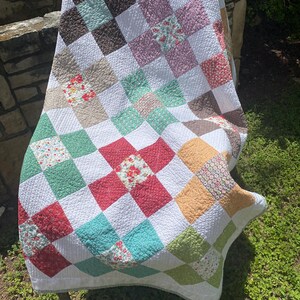 K&ouml;nnte beinhalten: Eine Patchwork-Decke mit einem Gitter aus abwechselnd wei&szlig;en Quadraten und farbig gemusterten Quadraten in Rot-, Gr&uuml;n-, Gelb-, Braun- und Rosat&ouml;nen. Der Stoff zeigt verschiedene Blumen- und geometrische Drucke mit durchgehend sichtbaren Steppn&auml;hten.