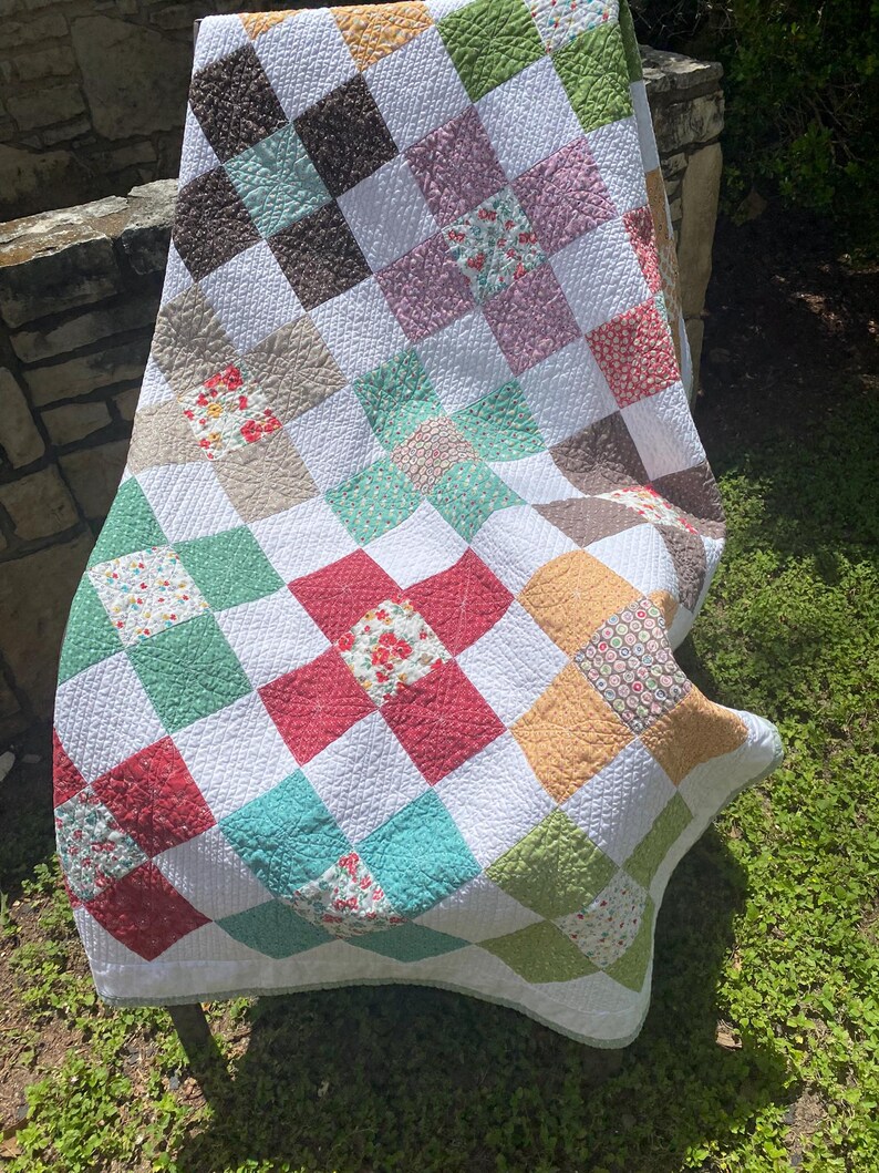 K&ouml;nnte beinhalten: Eine Patchwork-Decke mit einem Gitter aus abwechselnd wei&szlig;en Quadraten und farbig gemusterten Quadraten in Rot-, Gr&uuml;n-, Gelb-, Braun- und Rosat&ouml;nen. Der Stoff zeigt verschiedene Blumen- und geometrische Drucke mit durchgehend sichtbaren Steppn&auml;hten.