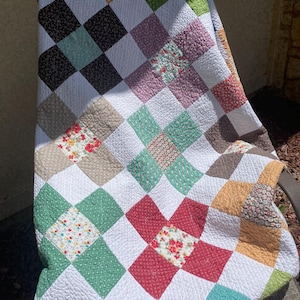 K&ouml;nnte beinhalten: Eine Patchwork-Decke mit einem sich wiederholenden Muster aus wei&szlig;en Quadraten, die sich mit Quadraten in verschiedenen Farben und Blumenmustern abwechseln, darunter Rot, Gr&uuml;n, Gelb, Braun und T&uuml;rkis. Der Stoff weist eine sichtbare Steppstruktur auf.
