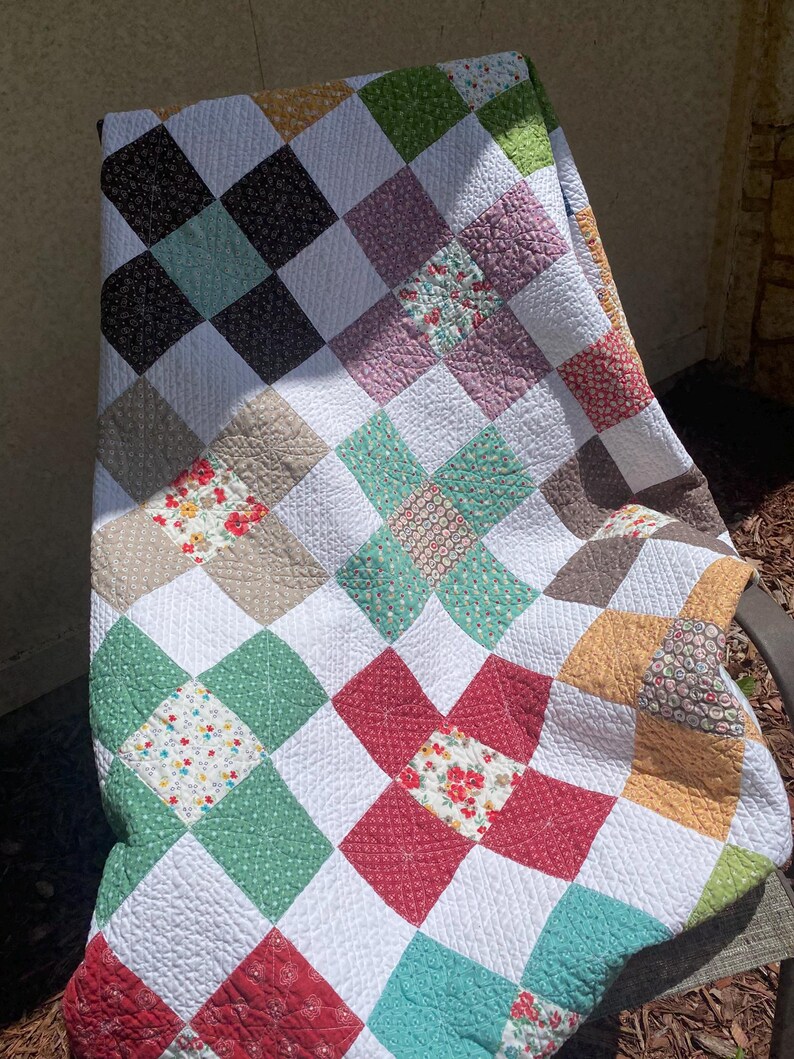 K&ouml;nnte beinhalten: Eine Patchwork-Decke mit einem sich wiederholenden Muster aus wei&szlig;en Quadraten, die sich mit Quadraten in verschiedenen Farben und Blumenmustern abwechseln, darunter Rot, Gr&uuml;n, Gelb, Braun und T&uuml;rkis. Der Stoff weist eine sichtbare Steppstruktur auf.