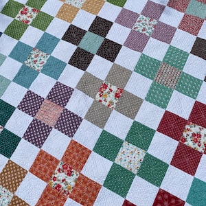 K&ouml;nnte beinhalten: Eine Patchwork-Decke mit einem sich wiederholenden Gittermuster aus wei&szlig;en Stoffquadraten, durchsetzt mit bunten floralen und geometrischen Druckquadraten in Rot-, Gr&uuml;n-, Blau-, Braun- und Gelbt&ouml;nen, die alle mit sichtbaren Steppn&auml;hten verbunden sind.