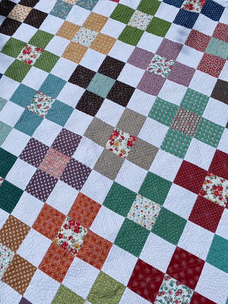 K&ouml;nnte beinhalten: Eine Patchwork-Decke mit einem sich wiederholenden Gittermuster aus wei&szlig;en Stoffquadraten, durchsetzt mit bunten floralen und geometrischen Druckquadraten in Rot-, Gr&uuml;n-, Blau-, Braun- und Gelbt&ouml;nen, die alle mit sichtbaren Steppn&auml;hten verbunden sind.