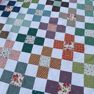 K&ouml;nnte beinhalten: Eine Patchwork-Decke mit einem sich wiederholenden Gittermuster aus wei&szlig;en Quadraten, die mit bunten Stoffquadraten durchsetzt sind. Die farbigen Quadrate zeigen verschiedene Blumen-, Punkte- und geometrische Drucke in Gr&uuml;n-, Rot-, Orange- und Violettt&ouml;nen.