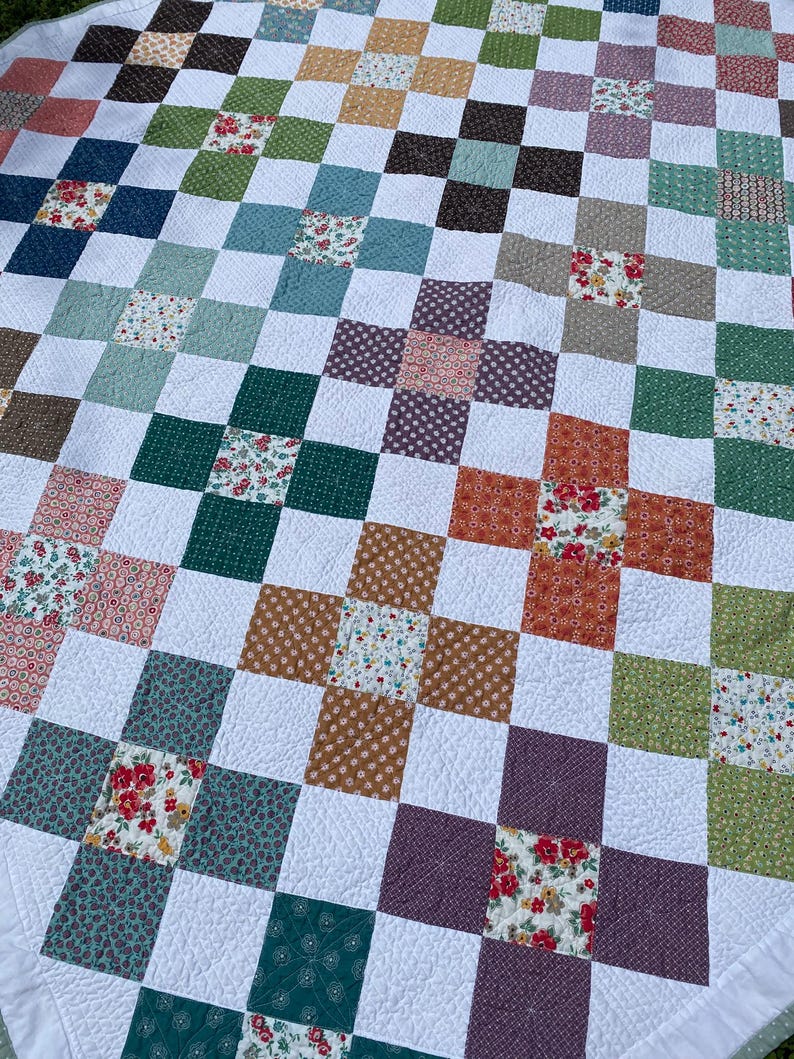 K&ouml;nnte beinhalten: Eine Patchwork-Decke mit einem sich wiederholenden Gittermuster aus wei&szlig;en Quadraten, die mit bunten Stoffquadraten durchsetzt sind. Die farbigen Quadrate zeigen verschiedene Blumen-, Punkte- und geometrische Drucke in Gr&uuml;n-, Rot-, Orange- und Violettt&ouml;nen.