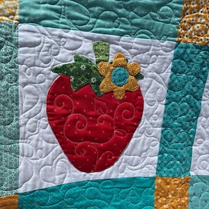 May include: A patchwork quilt featuring a central red strawberry appliqu&eacute; with a green stem and a yellow flower accent. The quilt is constructed from white fabric blocks with swirling heart quilting patterns, bordered by teal and yellow patterned fabric strips.