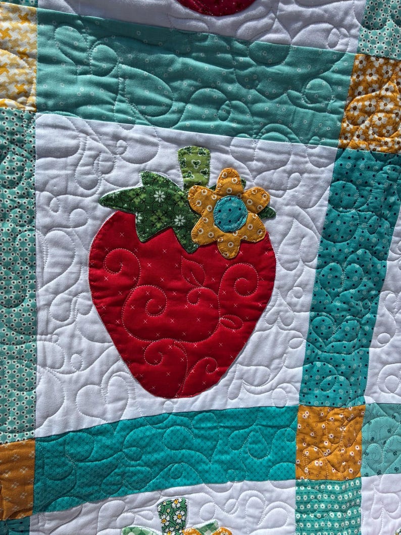 May include: A patchwork quilt featuring a central red strawberry appliqu&eacute; with a green stem and a yellow flower accent. The quilt is constructed from white fabric blocks with swirling heart quilting patterns, bordered by teal and yellow patterned fabric strips.