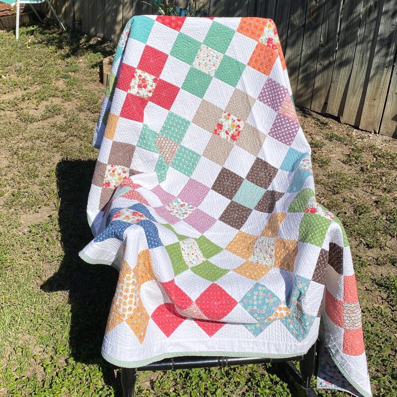 K&ouml;nnte beinhalten: Eine Patchwork-Decke mit einem Gitter aus abwechselnden wei&szlig;en Quadraten und bunten gemusterten Quadraten in Rot-, Gr&uuml;n-, Orange-, Blau- und Braunt&ouml;nen. Die Stoffquadrate sind durch sichtbare Steppn&auml;hte &uuml;ber das gesamte Textil hinweg verbunden.