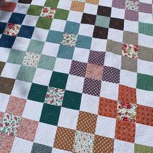 K&ouml;nnte beinhalten: Eine Patchwork-Decke mit einem sich wiederholenden Muster aus wei&szlig;en Quadraten, die mit bunten Bl&ouml;cken durchsetzt sind. Die Bl&ouml;cke bestehen aus verschiedenen Blumen- und geometrischen Drucken in Gr&uuml;n-, Blau-, Braun-, Orange-, Rosa- und Violettt&ouml;nen.
