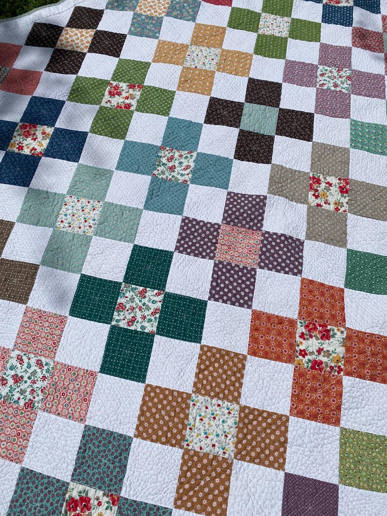 K&ouml;nnte beinhalten: Eine Patchwork-Decke mit einem sich wiederholenden Muster aus wei&szlig;en Quadraten, die mit bunten Bl&ouml;cken durchsetzt sind. Die Bl&ouml;cke bestehen aus verschiedenen Blumen- und geometrischen Drucken in Gr&uuml;n-, Blau-, Braun-, Orange-, Rosa- und Violettt&ouml;nen.