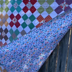 K&ouml;nnte beinhalten: Ein Patchwork-Quilt mit einer rautenf&ouml;rmigen Oberseite aus mehrfarbigen Stoffquadraten in Rot, Gr&uuml;n, Braun und Blumenmustern, die durch wei&szlig;en Stoff getrennt sind. Die R&uuml;ckseite zeigt einen wei&szlig;en Stoff mit einem wiederkehrenden Muster aus blauen V&ouml;geln.