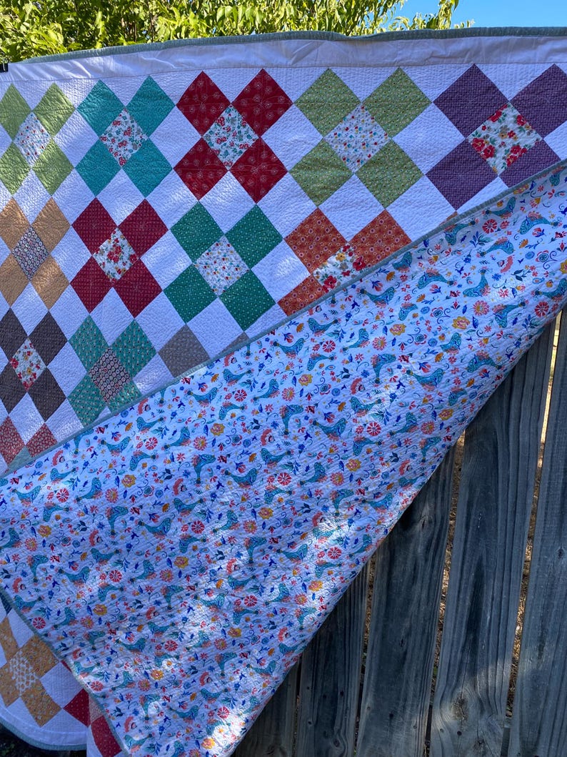 K&ouml;nnte beinhalten: Ein Patchwork-Quilt mit einer rautenf&ouml;rmigen Oberseite aus mehrfarbigen Stoffquadraten in Rot, Gr&uuml;n, Braun und Blumenmustern, die durch wei&szlig;en Stoff getrennt sind. Die R&uuml;ckseite zeigt einen wei&szlig;en Stoff mit einem wiederkehrenden Muster aus blauen V&ouml;geln.