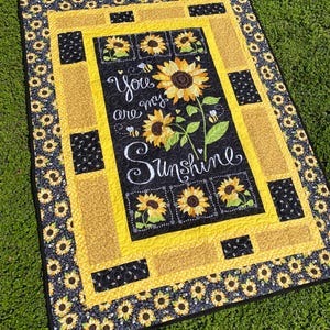 May include: A rectangular quilt featuring a sunflower design. The quilt has a black center with the words "You are my Sunshine" and images of sunflowers and bees. The border is yellow and black with sunflower patterns.
