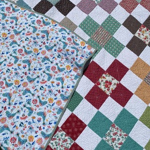 K&ouml;nnte beinhalten: Eine Steppdecke mit einem Patchwork-Design aus wei&szlig;en Quadraten, die sich mit Bl&ouml;cken aus floralen, geometrischen und einfarbigen Stoffquadraten in Rot, Gr&uuml;n, Braun und Gelb abwechseln. Die umgeschlagene Ecke zeigt einen wei&szlig;en Stoff mit Vogelmuster.