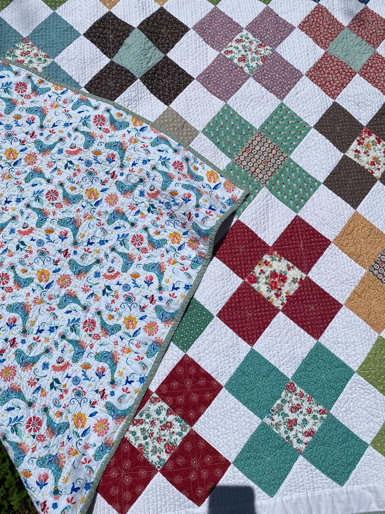 K&ouml;nnte beinhalten: Eine Steppdecke mit einem Patchwork-Design aus wei&szlig;en Quadraten, die sich mit Bl&ouml;cken aus floralen, geometrischen und einfarbigen Stoffquadraten in Rot, Gr&uuml;n, Braun und Gelb abwechseln. Die umgeschlagene Ecke zeigt einen wei&szlig;en Stoff mit Vogelmuster.