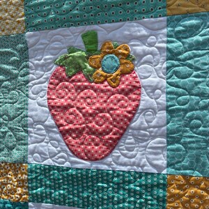 May include: A patchwork quilt featuring a central strawberry appliqu&eacute; with a pink polka dot pattern, a green stem, and a yellow flower. The quilt is constructed from white, teal, and yellow fabric squares with various patterns and swirling stitching.