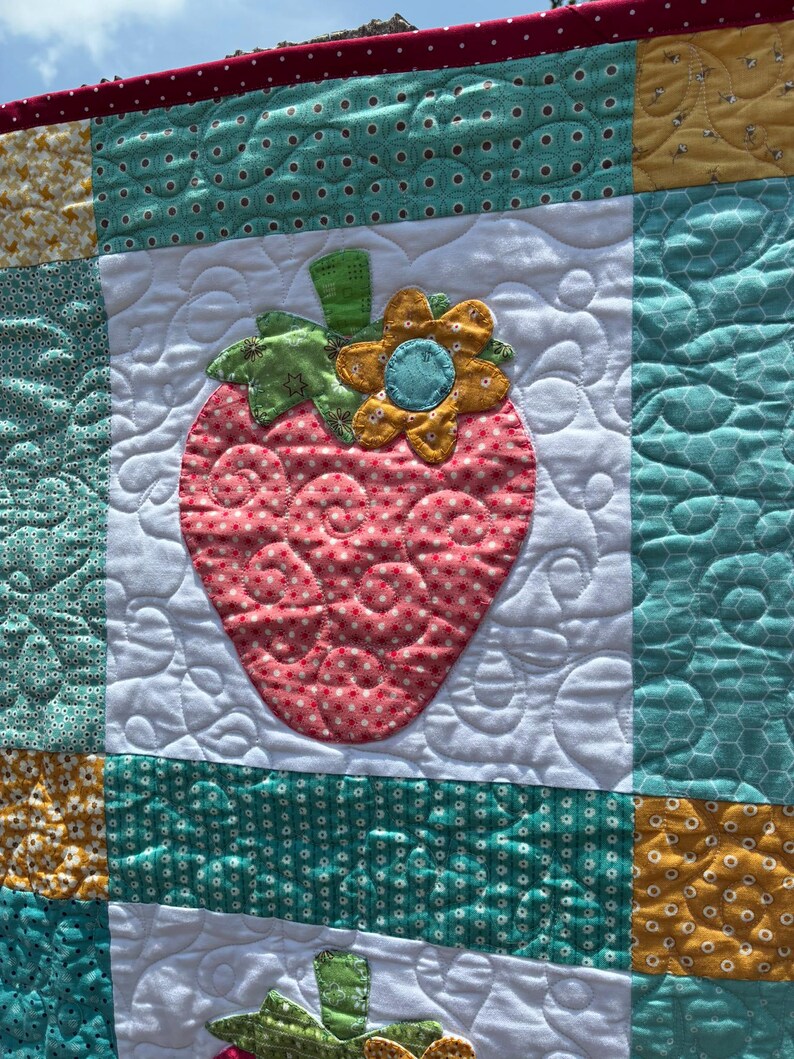 May include: A patchwork quilt featuring a central strawberry appliqu&eacute; with a pink polka dot pattern, a green stem, and a yellow flower. The quilt is constructed from white, teal, and yellow fabric squares with various patterns and swirling stitching.