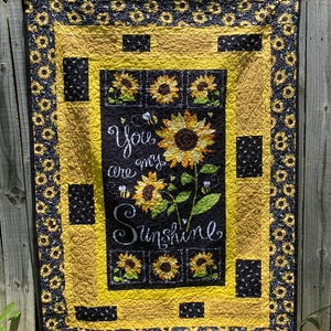 May include: A handmade quilt with a sunflower theme. The quilt features a black center panel with the words "You are my sunshine" and images of sunflowers and bees. The border is yellow and black with sunflower patterns.