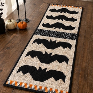 May include: A rectangular quilted table runner featuring a repeating pattern of black fabric bats appliqued onto a cream-colored background with a swirling texture. The runner is bordered by a black trim and a colorful orange, white, and black diamond pattern.