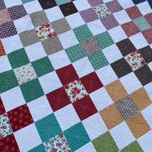 K&ouml;nnte beinhalten: Eine Patchwork-Decke mit einem sich wiederholenden Muster aus wei&szlig;en Quadraten, durchsetzt mit bunten Stoffquadraten in Rot-, Gr&uuml;n-, Blau-, Braun- und Gelbt&ouml;nen. Die Stoffquadrate zeigen verschiedene Blumen- und geometrische Drucke mit sichtbaren Steppn&auml;hten.