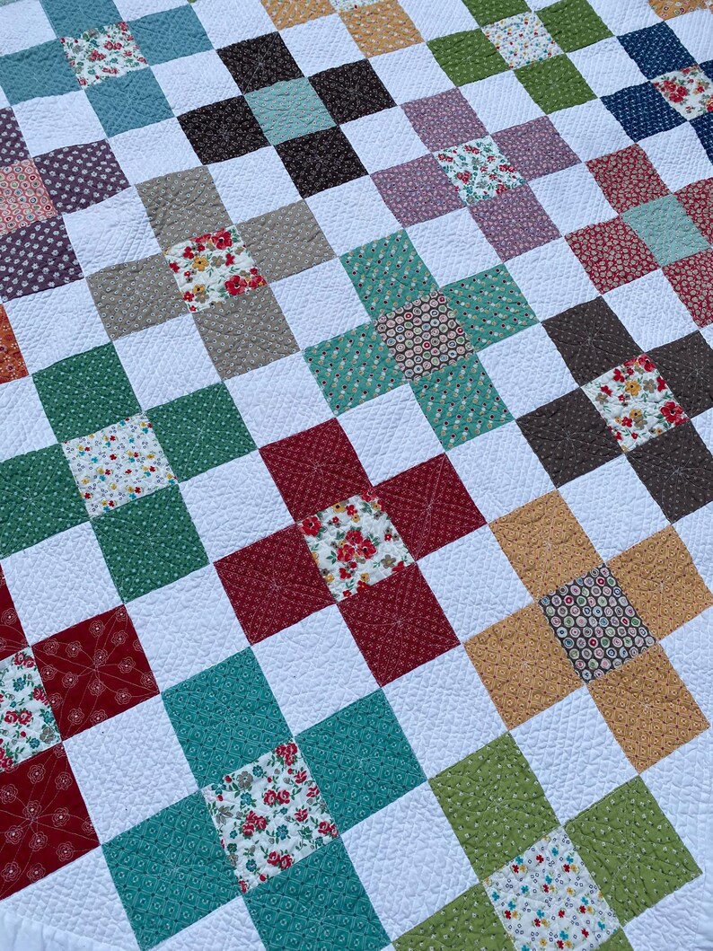 K&ouml;nnte beinhalten: Eine Patchwork-Decke mit einem sich wiederholenden Muster aus wei&szlig;en Quadraten, durchsetzt mit bunten Stoffquadraten in Rot-, Gr&uuml;n-, Blau-, Braun- und Gelbt&ouml;nen. Die Stoffquadrate zeigen verschiedene Blumen- und geometrische Drucke mit sichtbaren Steppn&auml;hten.