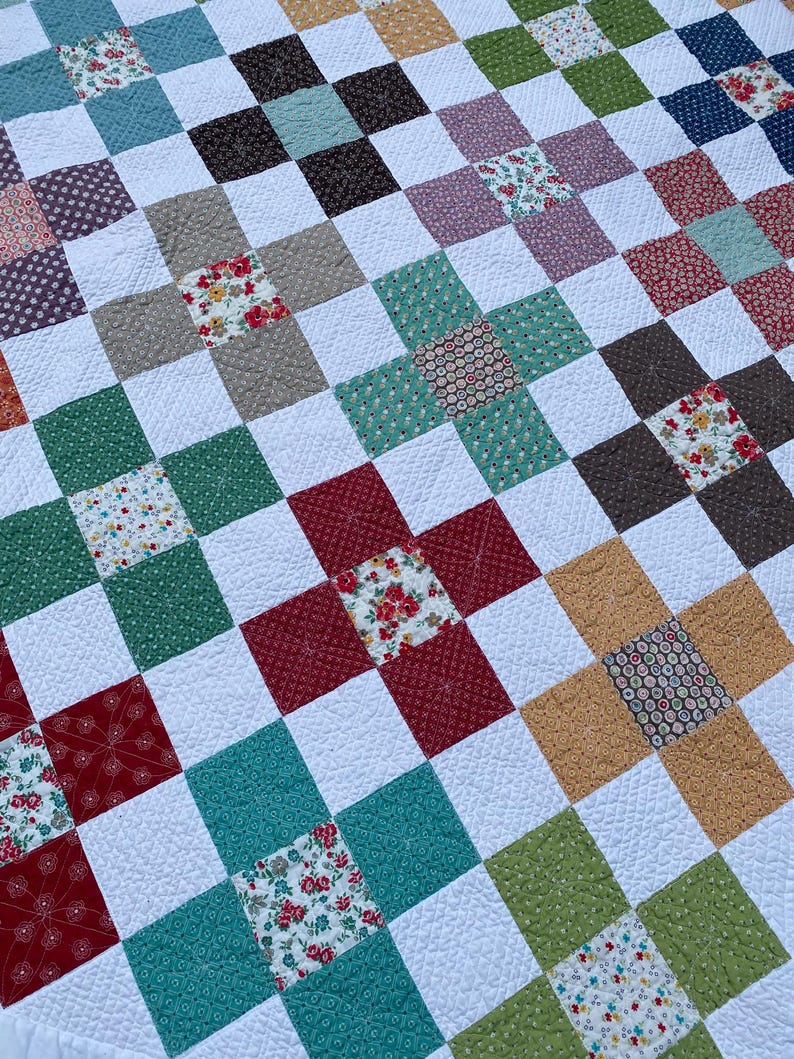 K&ouml;nnte beinhalten: Ein Patchwork-Quilt mit einem sich wiederholenden Gittermuster aus wei&szlig;en Quadraten, die mit bunten Quadraten durchsetzt sind. Die farbigen Quadrate zeigen verschiedene Blumenmuster, einfarbige Texturen sowie geometrische Muster in Rot, Gr&uuml;n, Blau und Gelb.