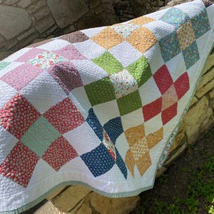 K&ouml;nnte beinhalten: Eine Patchwork-Decke mit einem Gitter aus quadratischen Stoffbl&ouml;cken in verschiedenen Farben wie Rot, Gr&uuml;n, Gelb, Blau und Braun, durchsetzt mit wei&szlig;en Quadraten. Die Decke hat eine texturierte, gesteppte Oberfl&auml;che und eine hellgr&uuml;ne Bord&uuml;re.