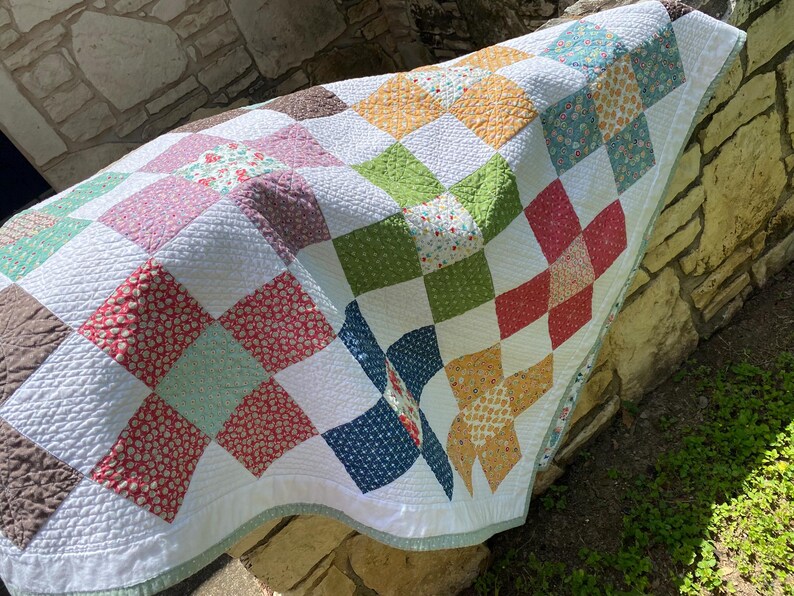 K&ouml;nnte beinhalten: Eine Patchwork-Decke mit einem Gitter aus quadratischen Stoffbl&ouml;cken in verschiedenen Farben wie Rot, Gr&uuml;n, Gelb, Blau und Braun, durchsetzt mit wei&szlig;en Quadraten. Die Decke hat eine texturierte, gesteppte Oberfl&auml;che und eine hellgr&uuml;ne Bord&uuml;re.
