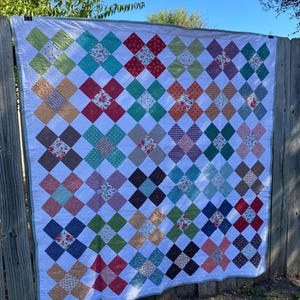 K&ouml;nnte beinhalten: Ein rechteckiger Quilt mit einem sich wiederholenden Muster aus X-f&ouml;rmigen Bl&ouml;cken, die aus verschiedenen bunten Stoffen mit Blumen-, Punkt- und Unimustern bestehen, getrennt durch wei&szlig;e Stoffquadrate und eingefasst mit einer hellgr&uuml;nen Bord&uuml;re.