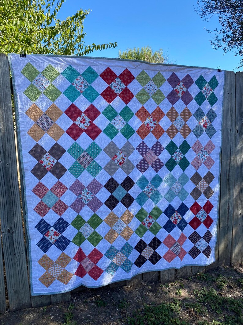 K&ouml;nnte beinhalten: Ein rechteckiger Quilt mit einem sich wiederholenden Muster aus X-f&ouml;rmigen Bl&ouml;cken, die aus verschiedenen bunten Stoffen mit Blumen-, Punkt- und Unimustern bestehen, getrennt durch wei&szlig;e Stoffquadrate und eingefasst mit einer hellgr&uuml;nen Bord&uuml;re.