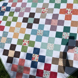 K&ouml;nnte beinhalten: Eine Patchwork-Decke mit einem sich wiederholenden Muster aus wei&szlig;en Quadraten, durchsetzt mit farbigen Quadraten in verschiedenen Blau-, Gr&uuml;n-, Rot-, Orange- und Braunt&ouml;nen, von denen viele kleine Blumenmuster und geometrische Designs aufweisen.