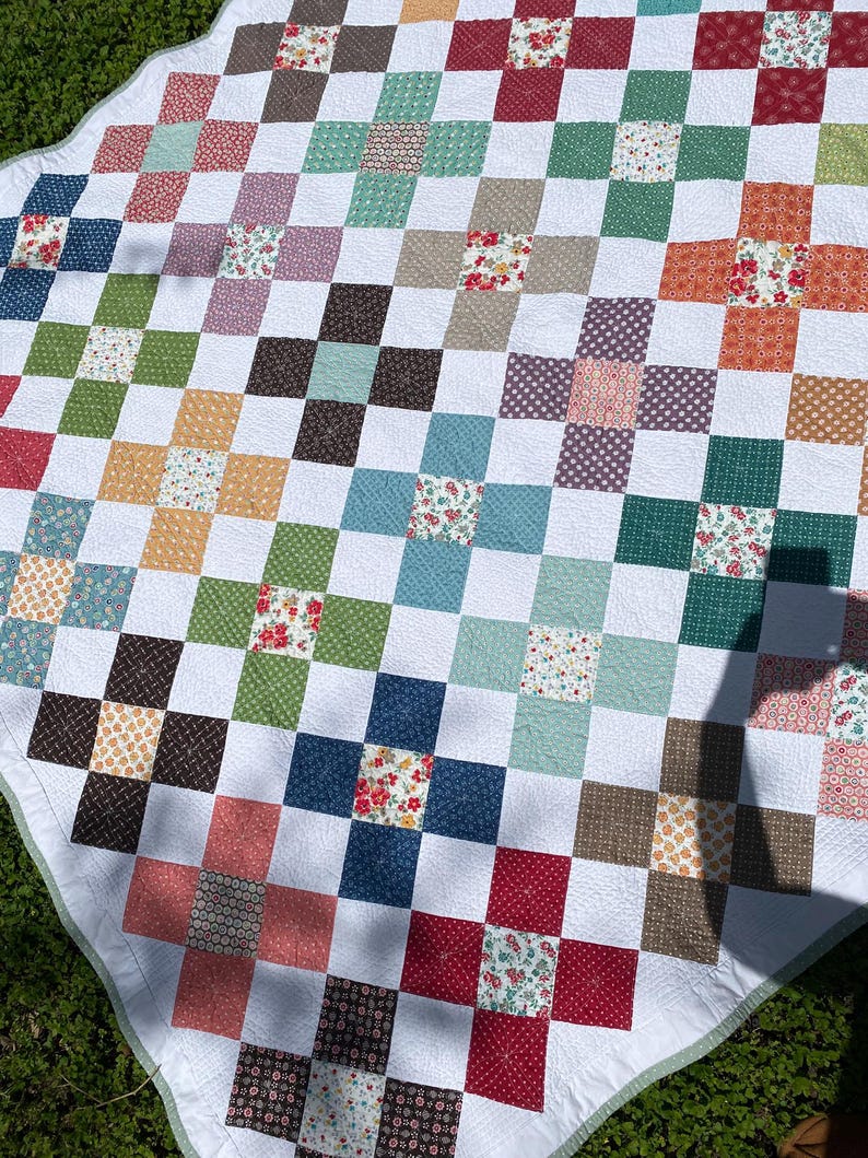 K&ouml;nnte beinhalten: Eine Patchwork-Decke mit einem sich wiederholenden Muster aus wei&szlig;en Quadraten, durchsetzt mit farbigen Quadraten in verschiedenen Blau-, Gr&uuml;n-, Rot-, Orange- und Braunt&ouml;nen, von denen viele kleine Blumenmuster und geometrische Designs aufweisen.