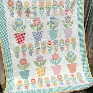 May include: A handmade quilt with a floral design. The quilt features gingham flowers in pots, with petals in yellow, red, blue, and purple. The quilt has a white background and a green and white checkered border.